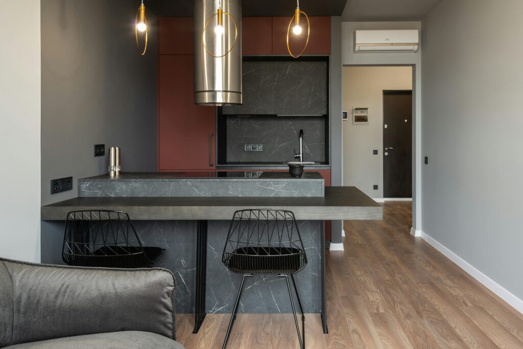 Dining zone furnished with counter and chairs in modern apartment with island style kitchen equipped with range hood and illuminated by hanging lamps