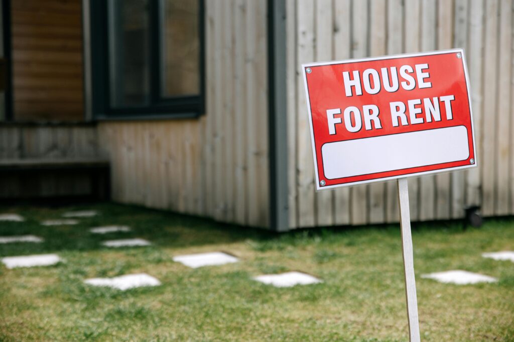 A modern wooden house with a prominent 'House for Rent' sign in the green yard.